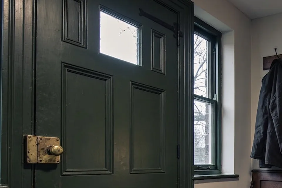 The inside of a deep green panelled front door in a pale plastered hall, a small clear pane in the top rail between two narrow panels, a black iron strap hinge across the top of the door, an old brass box lock with a round brass knob screwed to the face of the door at the left, and a sash window beside the door showing bare winter trees