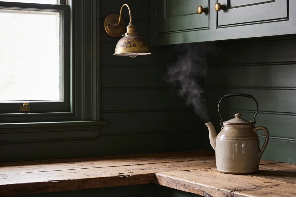A corner of a kitchen painted one deep green all over: horizontal boards on the walls, the window frame and sill, and the panelled cupboard doors above and below, with small brass knobs and a cup pull, an old brass wall lamp with a worn shade, a scrubbed wooden worktop, and a pale stoneware kettle steaming in the window light