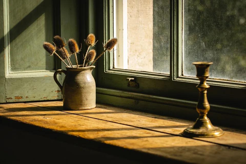 A deep-set cottage windowsill in low golden light — stoneware, dried seed heads, worn brass